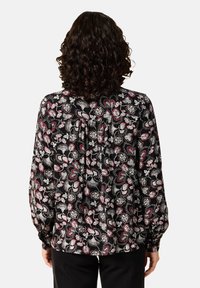 Person med lockigt mörkt hår bär en svart blus med vitt och rött blommönster, står vänd bort från kameran mot vit bakgrund.