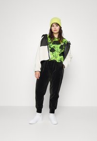 Čierne sametové joggery, neónovo zelený grafický top, biela obtiahnutá bunda a biele tenisky. Modelka má na hlave jasne žltú pletenú čiapku.