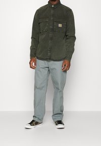 Ciemnozielona koszula z guzikami i dwoma kieszeniami na piersiach, zestawiona z jasnoszarymi spodniami i czarnymi trampkami w kamuflażu.