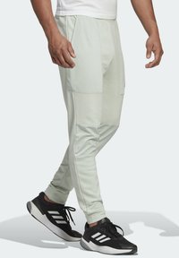 Helle grüne Jogginghosen mit seitlichen Taschen, schmal zulaufenden Beinen und einer glatten Textur, kombiniert mit schwarzen Sportschuhen mit weißen Streifen.