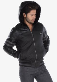 Black bomber jacket with textured faux snake skin sleeves, hood, zipper closure, and striped cuffs. Features logo patch on the chest.