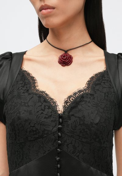 Zwarte kanten jurk met satijnen accenten, voorzien van een V-hals, decoratieve knopen en een zwarte koordketting met een bordeauxrode rozenhanger.