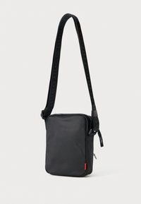 Sort nylon crossbody taske med rektangulær form, lynlåslukning og tekstureret skulderrem med præget branding. Rød logoaccent.
