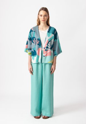 Cardigan lavorato a maglia in teal, rosa e blu navy con motivi floreali; abbinato a pantaloni a gamba larga azzurri; il modello si trova su uno sfondo neutro.