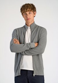 Grå zip-up cardigan med en texturerad stickad design, långa ärmar och ribbade manschetter. Buren över en vit t-shirt med hög krage.