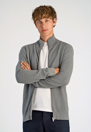 Grijze ritssluiting cardigan met een textuur gebreid ontwerp, lange mouwen en ribgebreide manchetten. Gedragen over een witte T-shirt met een hoge kraag.
