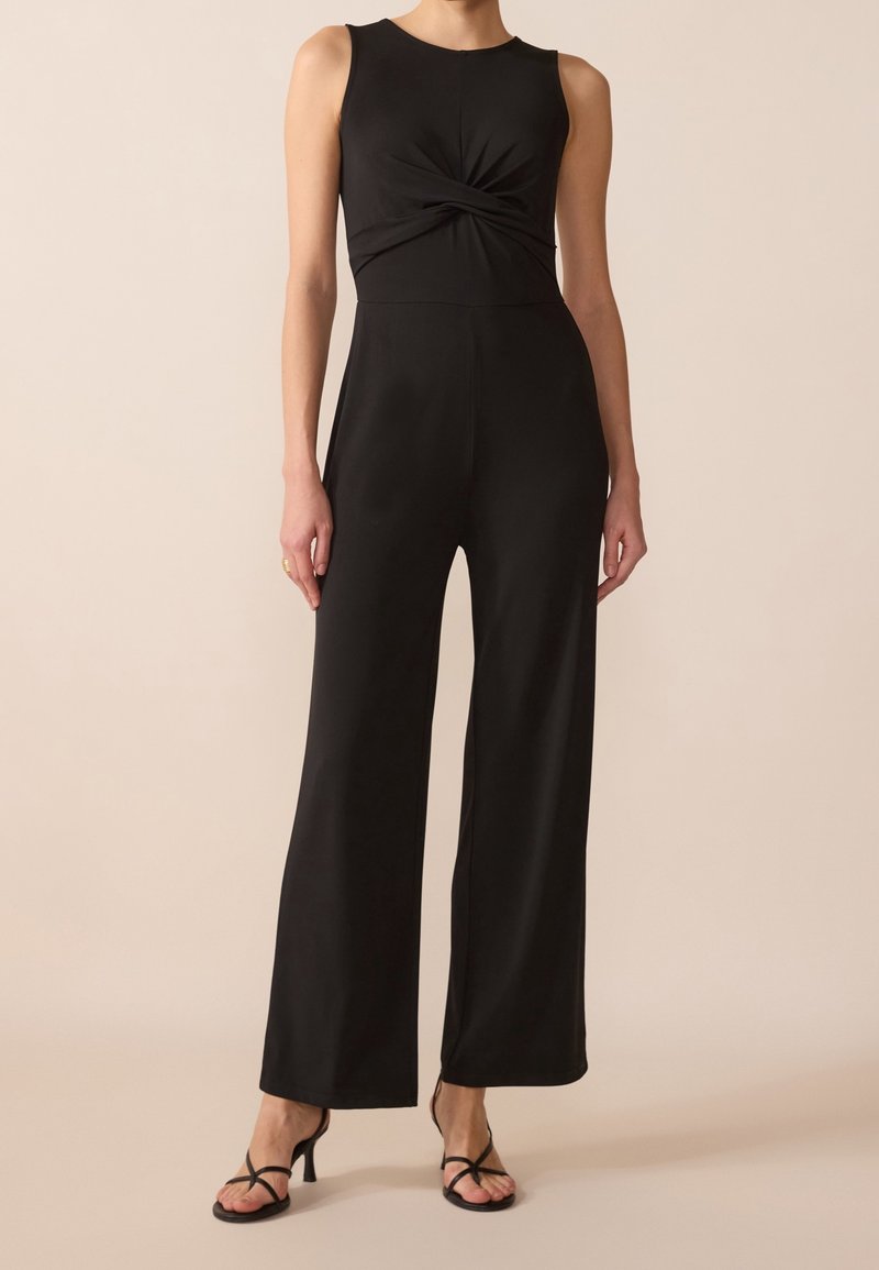 Femme portant une combinaison noire sans manches avec un détail de nœud torsadé à la taille et un pantalon large, associée à des talons noirs à brides.