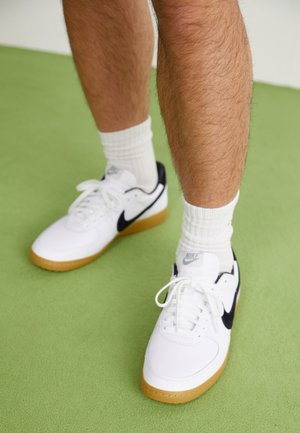 Sapatilhas Nike brancas com detalhes pretos e solas de borracha. Materiais em malha e couro. Usadas com meias brancas de canelado. Fundo verde.