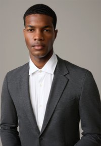 Garnitur w kolorze węgla z klapą typu notch i zapięciem na jeden guzik, zestawiony z białą koszulą z kołnierzykiem. Gładka tekstura materiału, minimalne detale.