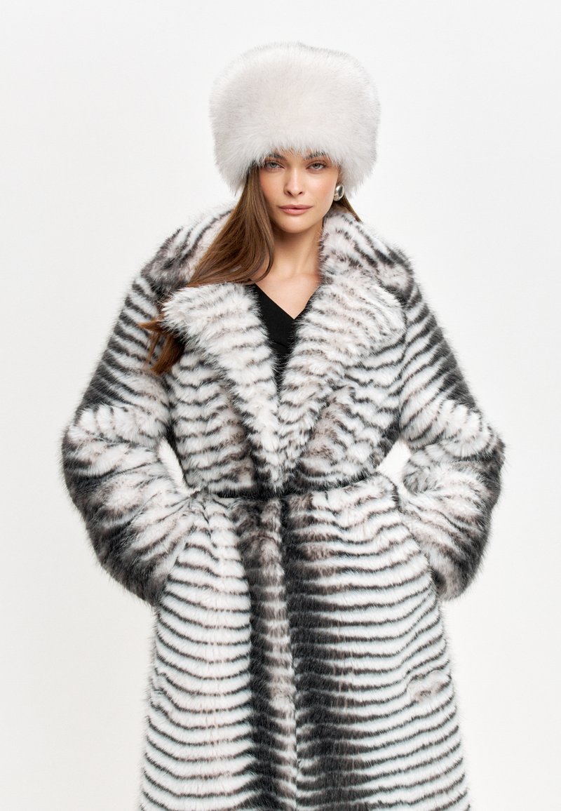 Cappotto in pelliccia sintetica con strisce bianche e nere, colletto ampio e cintura legata in vita. Abbinato a un cappello bianco soffice. Finitura liscia e texturizzata.