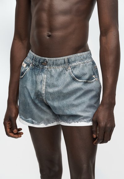 Γυμνός κορμός και χέρια ενός ατόμου που φοράει ξεθωριασμένα τζιν σορτς με φθαρμένες άκρες και λευκή επένδυση από κάτω, στέκεται μπροστά σε ένα απλό φόντο.