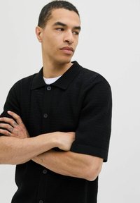 Black short-sleeve knit shirt with a collared neckline, ribbed texture, and five buttons down the front. Smooth fabric with subtle pattern.