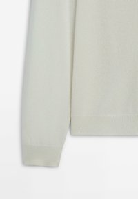 Poignet et ourlet d'un pull en maille crème à manches longues, présentés sur un fond blanc uni.