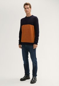 Gebreide trui met een marineblauw bovenstuk en een oranje onderstuk, voorzien van een textuurpatroon. Gecombineerd met donkerblauwe jeans en zwarte laarzen.