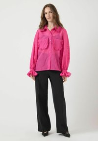 Vrouw gekleed in een doorschijnende felroze blouse met gerimpelde zakken en manchetten, gecombineerd met losse zwarte broek en zwarte puntige schoenen.