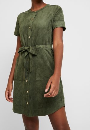 Kobieta ubrana w oliwkowo-zieloną sukienkę z krótkim rękawem, zapinaną na guziki z przodu, z paskiem do wiązania i dużymi przednimi kieszeniami.