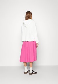 Bílá zkrácená mikina s kapucí, kombinovaná s růžovou sukní do kolen, obutá s černými teniskami a bílými pruhovanými ponožkami.