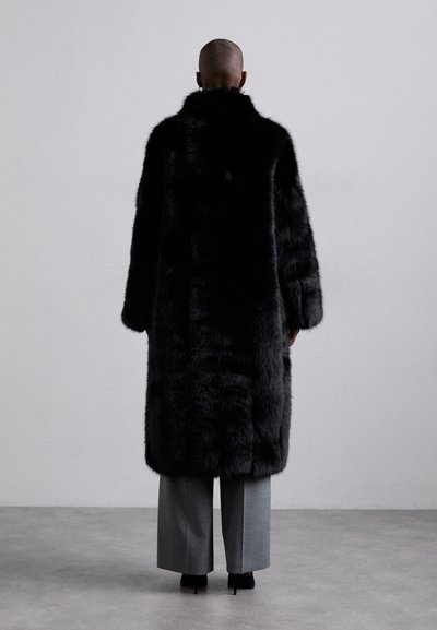 Schwarzer Kunstfellmantel mit hohem Kragen und weiten Ärmeln, kombiniert mit grauen weiten Hosen und High Heels, von hinten betrachtet.
