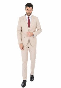 Completo beige chiaro con un design aderente, giacca monopetto e pantaloni coordinati. Abbinato a una camicia bianca e una cravatta rossa. Scarpe nere.