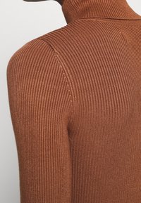 Gros plan sur une personne portant un pull à col roulé en laine marron côtelée, mettant l'accent sur la zone de l'épaule et du cou.