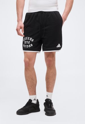 Mężczyzna w czarnych szortach Adidas z napisem „QUEENS NYC ADIDAS”, białych skarpetkach Adidas i czarnych butach sportowych Adidas, stoi na jednolitym tle.