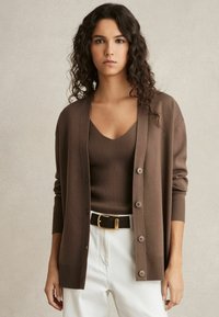Cardigan marrone realizzato in materiale morbido, con scollatura a V profonda, chiusura con bottoni e polsini a coste, indossato sopra un top coordinato e pantaloni bianchi.