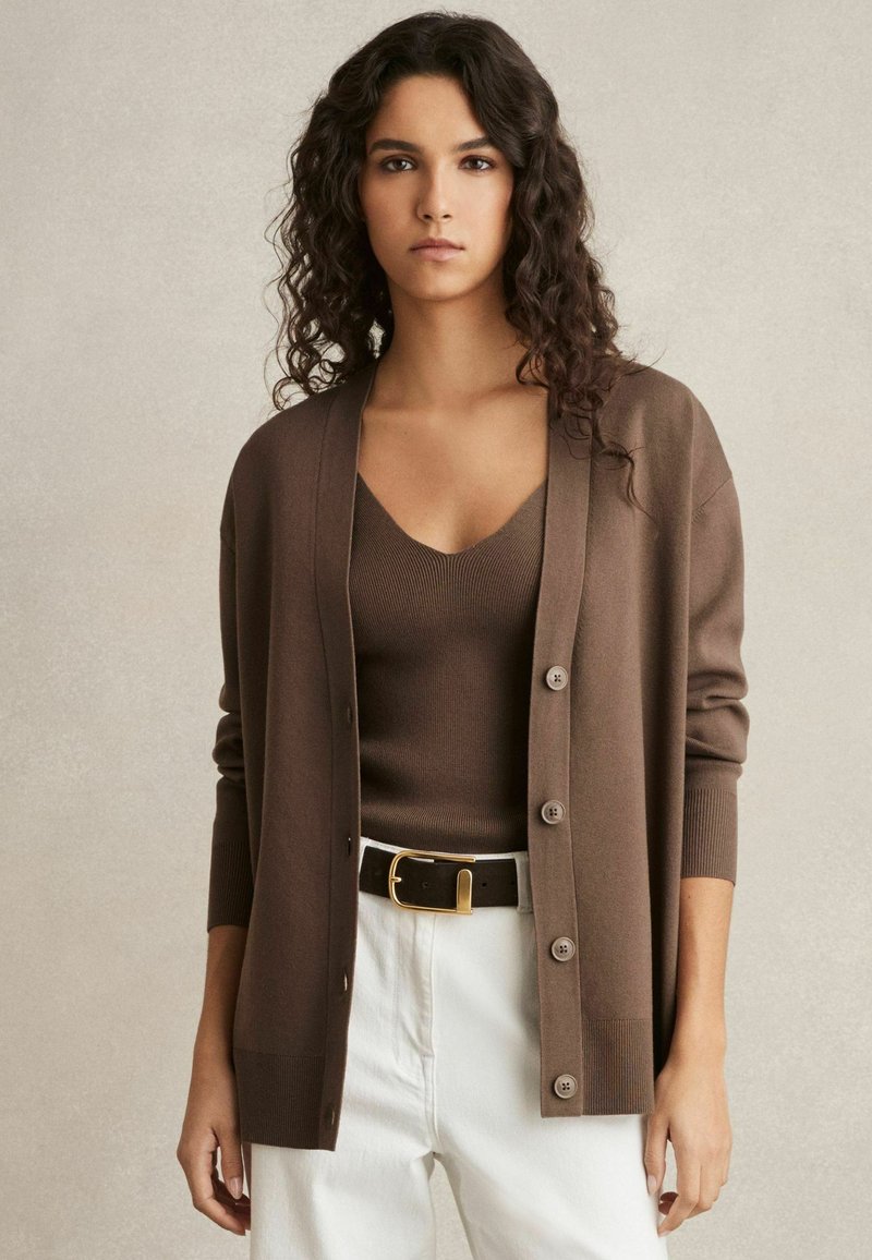 Brun cardigan i mjukt material, med djup V-ringning, knappstängning och ribbade ärmslut, bärs över en matchande topp och vita byxor.