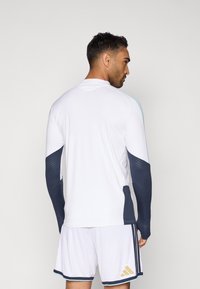 Άνδρας που φορά λευκά αθλητικά ρούχα με σκούρους γκρι μανίκες και σορτς που διαθέτει χρυσό λογότυπο, βλέπεται από πίσω σε απλό φόντο.