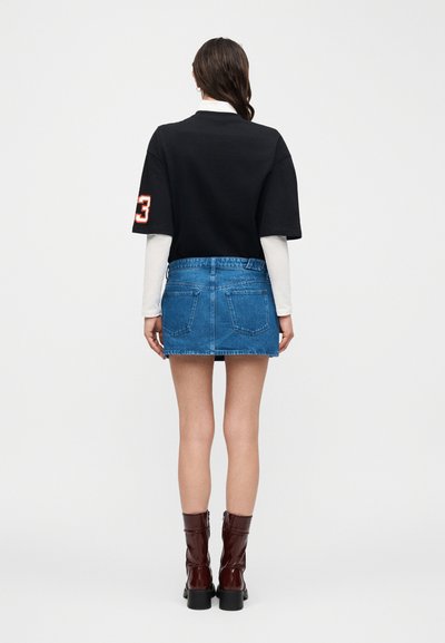 Γυναίκα που στέκεται με την πλάτη προς τον θεατή, φοράει μαύρο κοντό τοπ με κοντά μανίκια πάνω από λευκά μακριά μανίκια, μπλε μίνι φούστα από τζιν και καφέ μποτάκια μέχρι τον αστράγαλο.