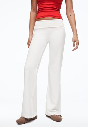 Woman wearing white wide-leg pants, red sleeveless top, and brown shoes, standing against a plain white background.