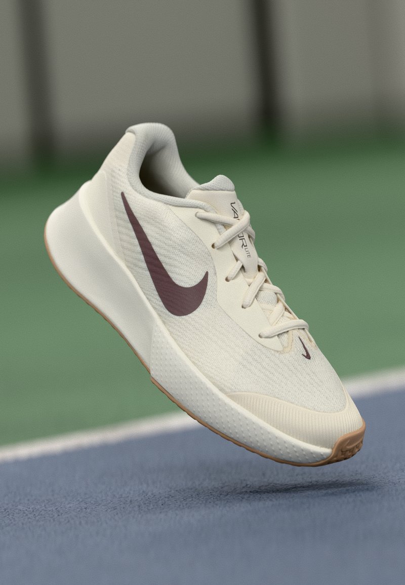 Beżowy but biegowy Nike z fioletowym logotypem Swoosh, białymi sznurówkami i gumową podeszwą unoszący się nad niebieską powierzchnią kortu tenisowego.