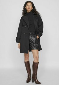 Svart trenchcoat med dubbelknäppt design, i kombination med en svart polotröja, shorts i fusk-läder och bruna knähöga stövlar.