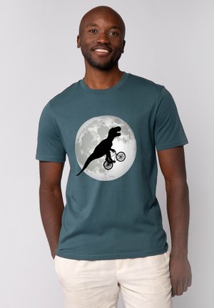 Homme souriant portant une chemise bleu sarcelle avec un graphique d'un T-Rex faisant du vélo devant une pleine lune.