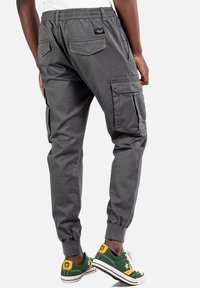 Graue Cargo-Hose mit elastischem Bund, seitlichen und hinteren Taschen. Taillierte Form und umgekrempelte Knöchel. Kombiniert mit grünen Turnschuhen mit gelben Akzenten.