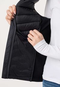 Ruky držia rozopnutú čiernu prešívanú bundu, ktorá odhaľuje jej hladkú vnútornú podšívku, oblečenú na bielej dlhorukávovej košeli a modrých džínsach.