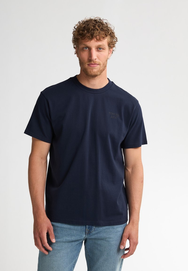 Donkerblauw katoenen T-shirt met een ronde hals en korte mouwen, voorzien van een subtiel logo aan de voorkant en een ontspannen pasvorm. Gecombineerd met lichtblauwe spijkerbroek.