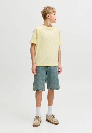 Niño con camiseta amarilla de manga corta, pantalones cortos vaqueros hasta la rodilla, calcetines blancos y zapatos beige con cordones, de pie mirando hacia un lado.