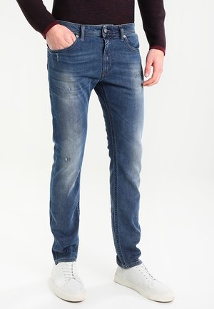 Mann trägt ausgewaschene blaue Jeans mit leichten Abnutzungsspuren und weiße Turnschuhe, Hand in der Tasche, steht vor einfachem Hintergrund.