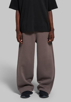 Personne portant un pantalon taupe à jambes larges, des baskets noires et une chemise noire ample à manches courtes, debout devant un fond gris.