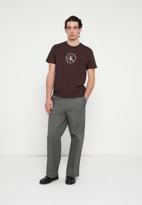 Hnedé bavlnené tričko s bielym kruhovým logom na prednej strane, v kombinácii s voľnými sivými nohavicami a čiernymi topánkami.