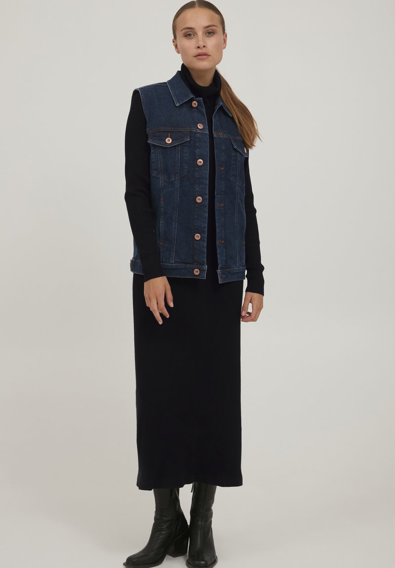 Gilet in denim blu scuro, con colletto, tasche frontali e bottoni in rame, abbinato a un vestito nero a maniche lunghe e stivali neri alla caviglia.