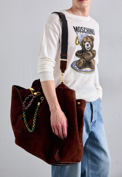Geantă mare din suede maro cu accesorii aurii, având mânere împletite în verde, galben și negru. Stilizată cu un pulover crem și pantaloni din denim.