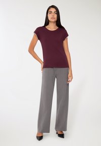 Kobieta stoi z ręką na biodrze, ubrana w krótką maroonową bluzkę, szerokie szare spodnie i czarne szpilki.