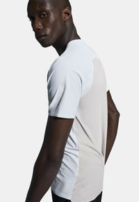 Vit sportt-shirt med texturerad tyg, korta ärmar och ett kontrasterande ljusgrått sidopanel. Designad för komfort och rörlighet.