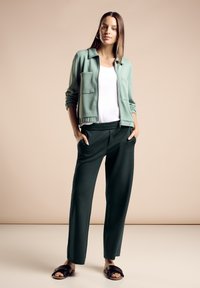 Kobieta stojąca z rękami w kieszeniach, ubrana w jasnooliwkową kurtkę na zamek, biały top, ciemnozielone spodnie i czarne klapki na beżowym tle.