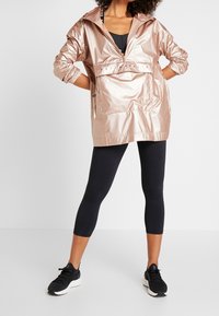 Metaliczna różowo-złota kurtka z kapturem, z przednią kieszenią na zamek i elastycznymi mankietami, zestawiona z czarnymi legginsami i sneakersami. Gładka, błyszcząca tekstura.