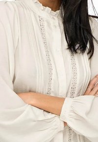 Weiße Bluse aus weichem Stoff mit hochrippigem Kragen, vertikalen Falten und Spitzenverzierungen entlang der Knopfleiste und Ärmel.