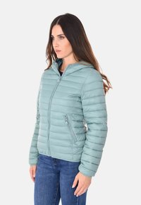 Giacca a vento leggera color teal con design a righe orizzontali, tasche con zip e cappuccio. Presenta una silhouette aderente e una texture liscia.