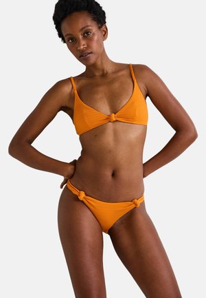 Femme debout les mains sur les hanches, portant un bikini orange texturé avec des détails de nœuds en haut et en bas, sur un fond clair uni.