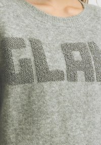 Siva pletena pulover s izstopajočim napisom "GLAM" v metalni niti. Tekstura se zdi mehka in puhasta z okroglim izrezom.